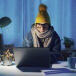 A woman works from home at her computer while wearing winter close.