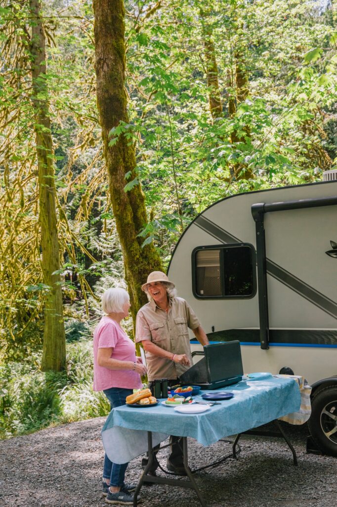 Two women cooking hot dogs at table in front of a camper at a recreation site.